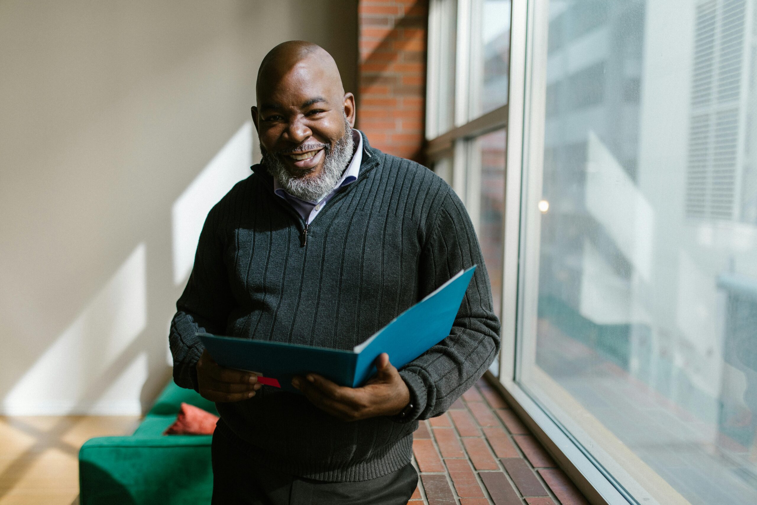 Cheerful man holding a folder in a sunlit room, smiling warmly.